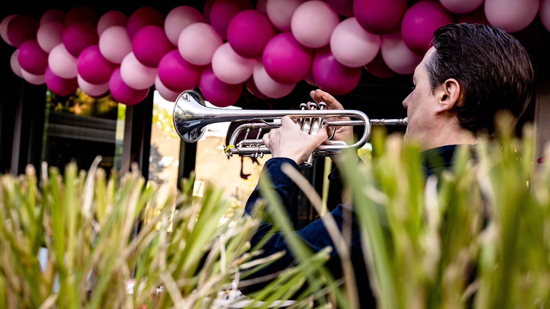Das Bild zeigt einen Trompeter, der bei einer Outdoor-Veranstaltung spielt. Im Hintergrund hängt eine dekorative Girlande aus rosa und pinken Luftballons, die eine festliche Atmosphäre schafft. Grüne Pflanzen im Vordergrund sorgen für eine natürliche Rahmung des Motivs. Die Szene vermittelt eine lebendige und elegante Stimmung, ideal für besondere Anlässe wie Feiern oder Events.
