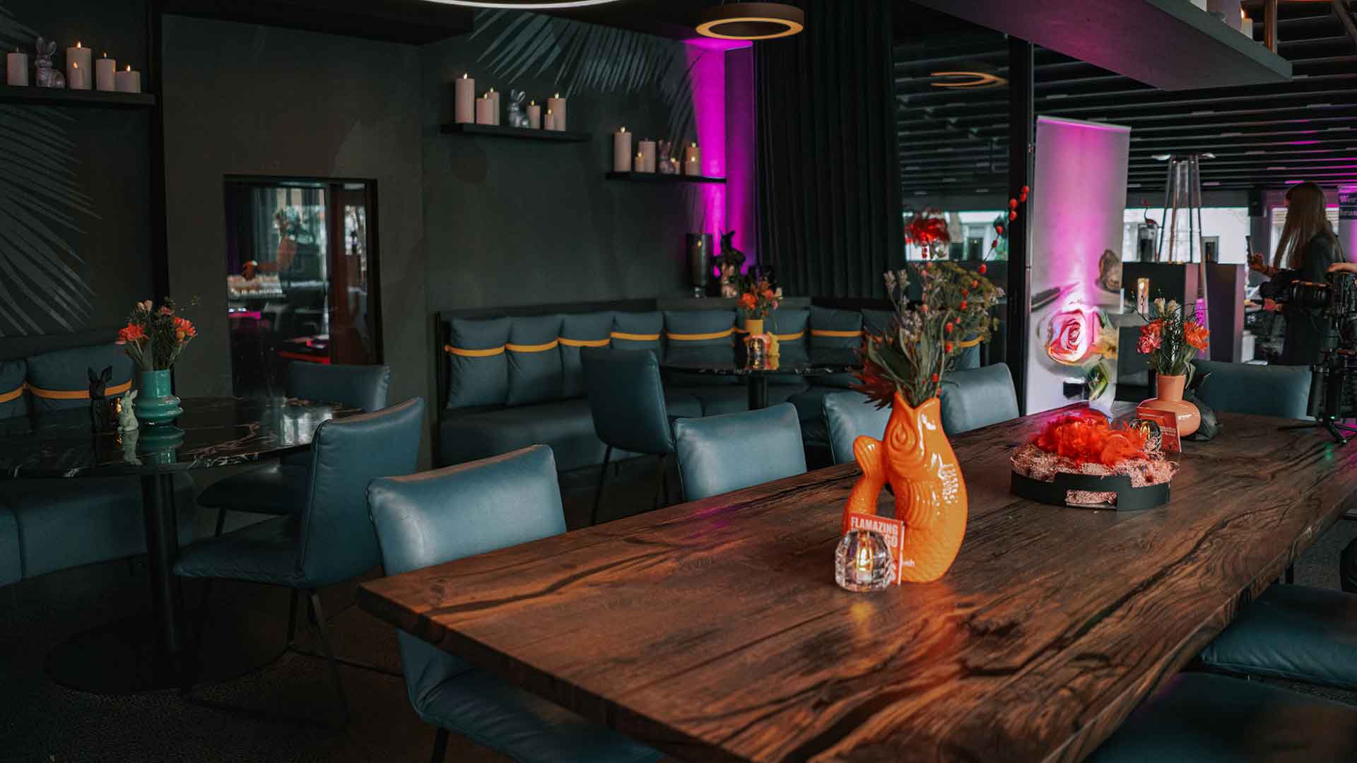 Stilvoll eingerichteter Restaurantbereich im Six Zero Kitchen & Bar mit modernen, dunklen Wänden, eleganten blauen Polstermöbeln und einem großen Holztisch. Die Tischdekoration umfasst farbenfrohe Vasen in Orange und Blumenschmuck. Im Hintergrund sind Kerzen und dezente Beleuchtung zu sehen.